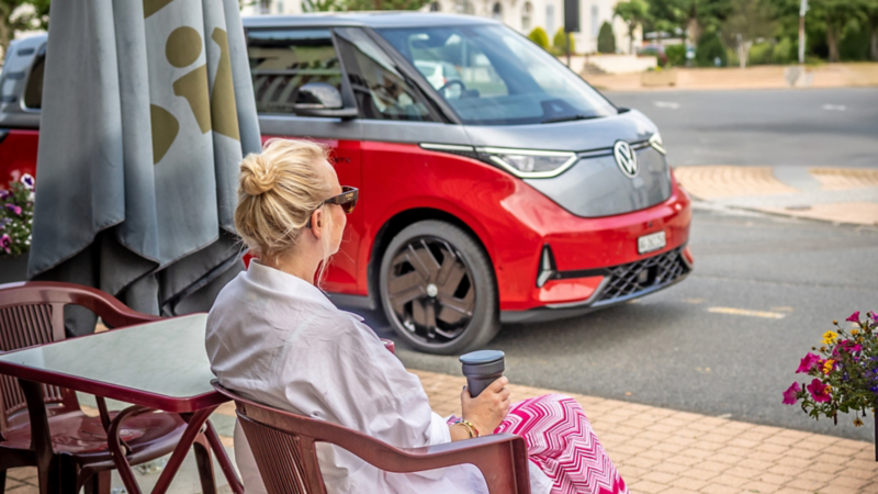 Eine Frau sitzt an einem Café-Tisch und blickt auf einen parkierten VW ID. Buzz vor ihr.