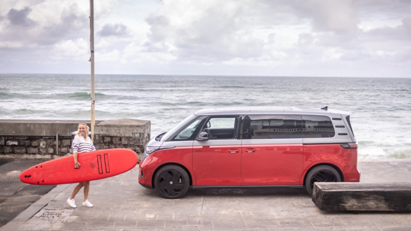 Eine Frau trägt ein rotes Surfboard neben einem VW ID. Buzz, direkt am Meer.