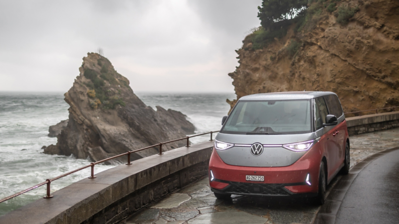 Ein VW ID. Buzz steht auf einer Küstenstrasse mit starken Wellen und Felsformationen im Hintergrund.