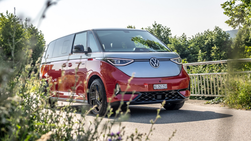 Der VW ID. Buzz fährt auf einer Landstrasse, mit Gräsern und Pflanzen im Vordergrund.