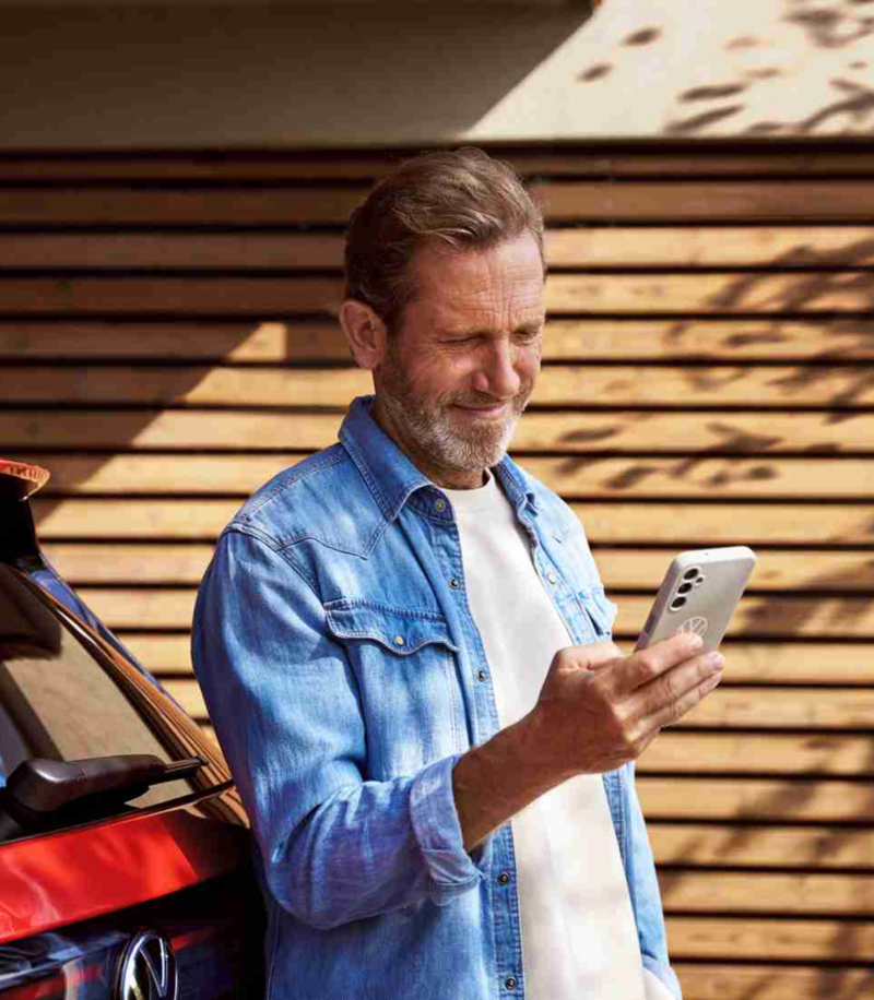 Middle-aged man with a beard in a denim shirt, standing next to a red car while looking at his smartphone