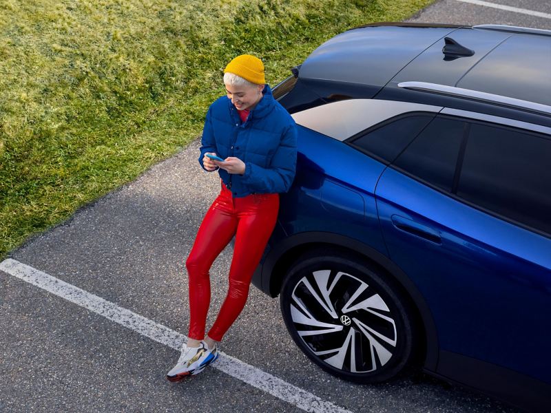 Une femme s’appuie contre sa Volkswagen tout électrique et cherche des informations sur la garantie mobilité à l’aide de son smartphone