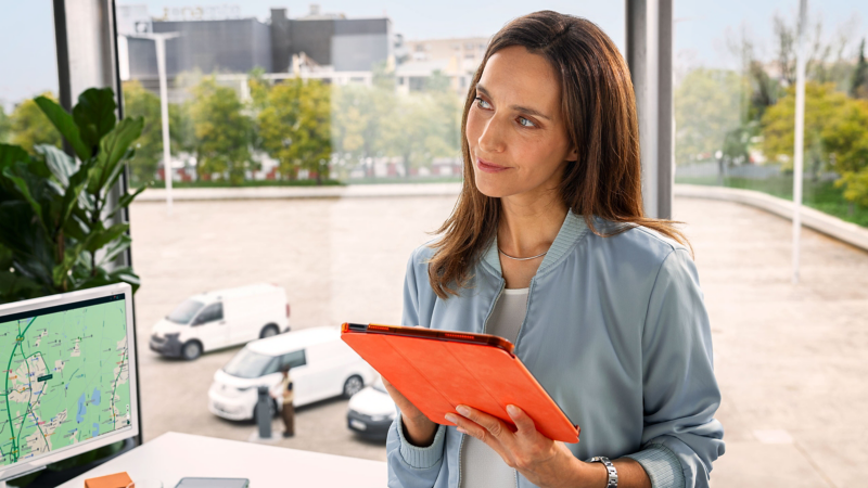 Eine Flottenmanagerin mit Tablet in der Hand im Büro, durch das Fenster sieht man VW Modelle auf dem Hof stehen.