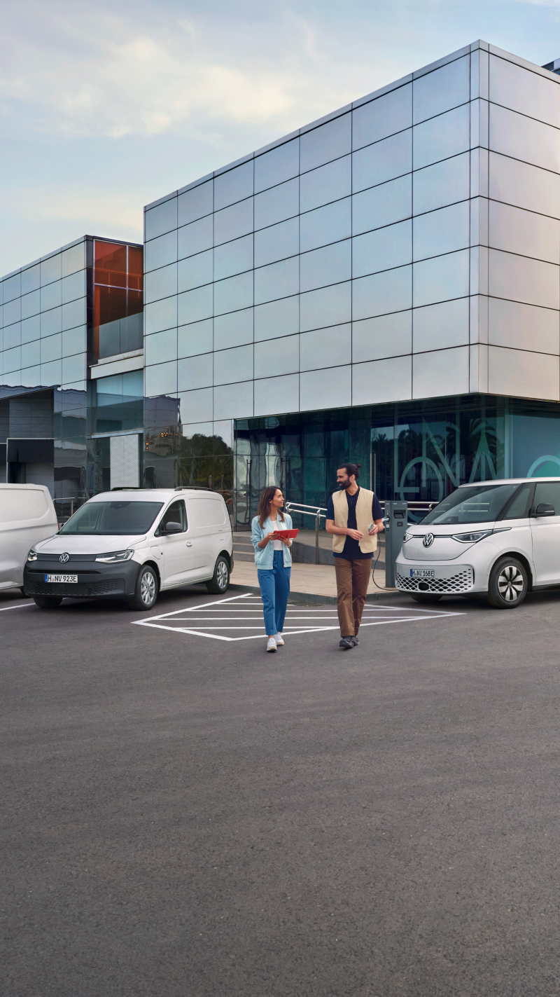 Ein Mann und eine Frau laufen auf einem Parkplatz auf dem Volkswagen Nutzfahrzeuge parken.