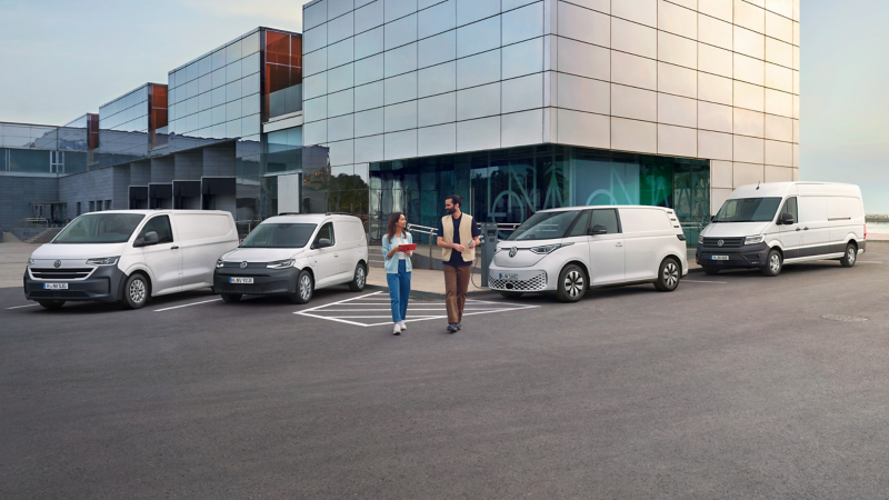Ein Mann und eine Frau laufen auf einem Parkplatz auf dem Volkswagen Nutzfahrzeuge parken.