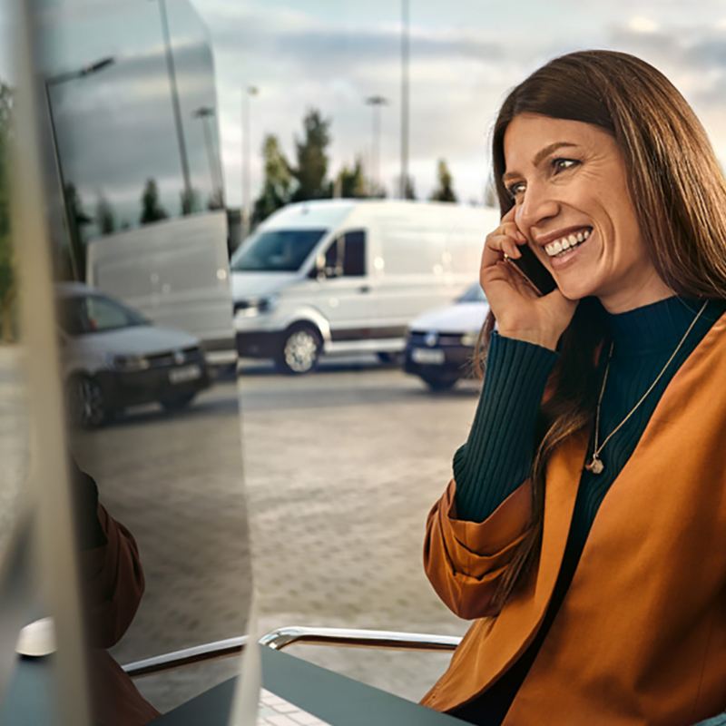 Una donna con un computer sta parlando al telefono in un parcheggio, alle sue spalle dei veicoli commerciali Volkswagen.