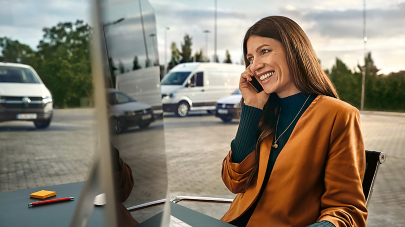 Una donna con un computer sta parlando al telefono in un parcheggio, alle sue spalle dei veicoli commerciali Volkswagen.