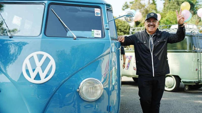 Hombre hace señal de amor y paz junto a Combi azul Volkswagen en evento de clásicos.