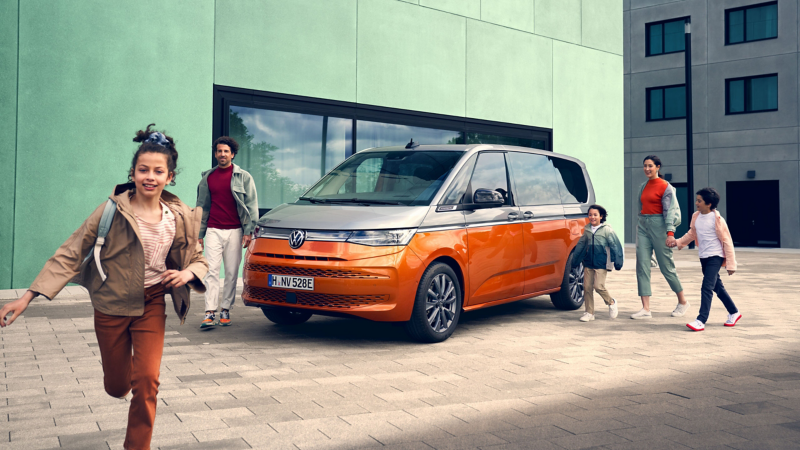 Vor einer modernen Architekturkulisse läuft ein Mädchen lachend im Vordergrund, während im Hintergrund eine Familie zu ihrem zweifarbigen, orange-silbernen VW Multivan geht.