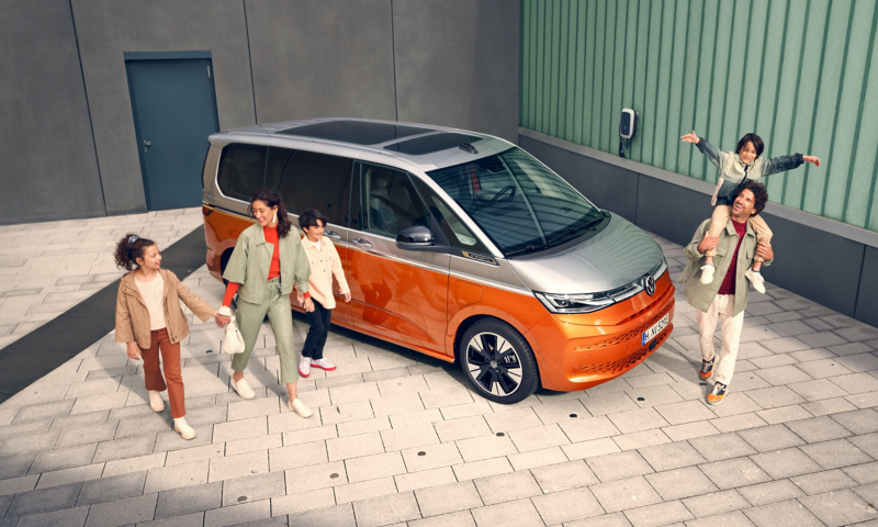 Una familia de cuatro (dos adultos y dos niños, uno de ellos sobre los hombros de un adulto) caminando alrededor de una furgoneta Volkswagen Multivan bicolor (naranja y plateada) estacionada en un espacio exterior moderno. La imagen resalta la capacidad familiar y la versatilidad del vehículo.