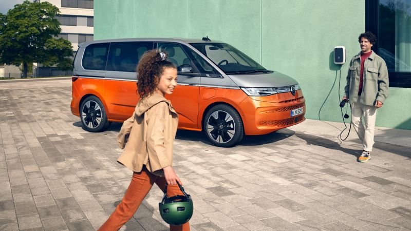 Un homme avec un câble de recharge à la main et une fille avec un casque devant le VW Multivan Energetic.