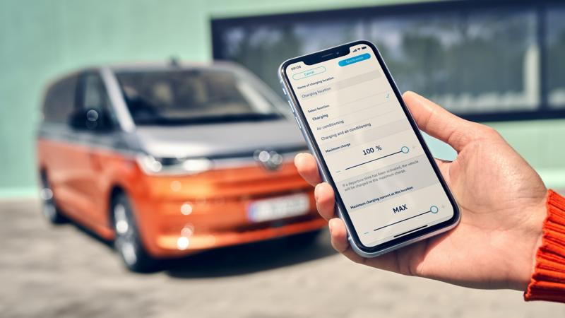 One hand holds a smartphone. In the background, the VW Multivan Energetic can be seen in a blur.