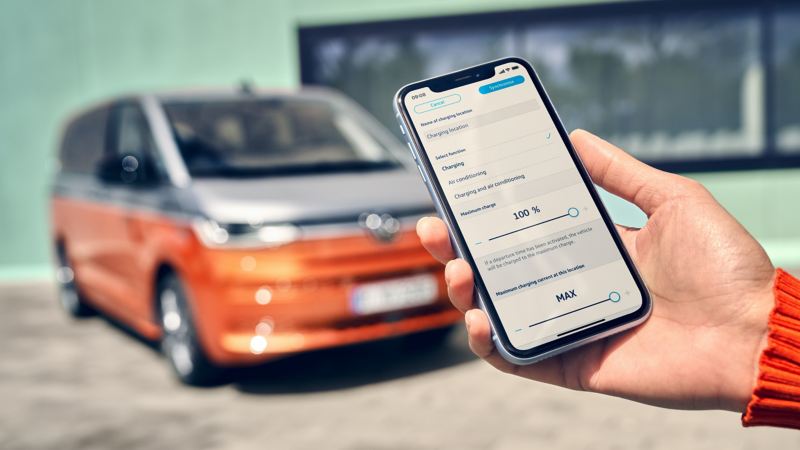 En smartphone holdes oppe foran en orange og grå VW Multivan