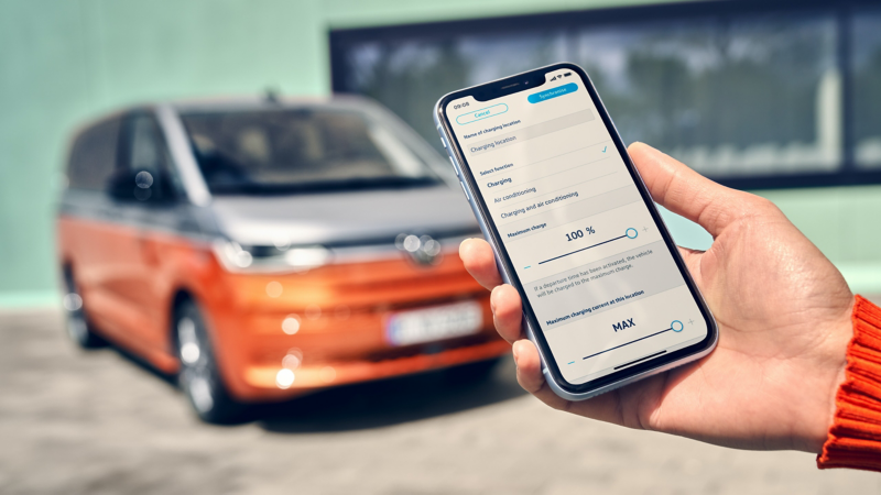 A hand holds a smartphone. In the background, the VW Multivan Energetic can be seen in a blur.