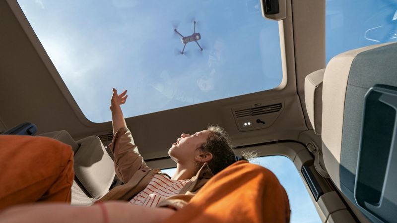 Dziewczynka wyciąga rękę w stronę szklanego dachu panoramicznego i patrzy na lecący nad autem dron.