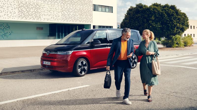 Far och dotter lämnar en parkerad röd-svart VW Multivan eHybrid minibuss och går över gatan