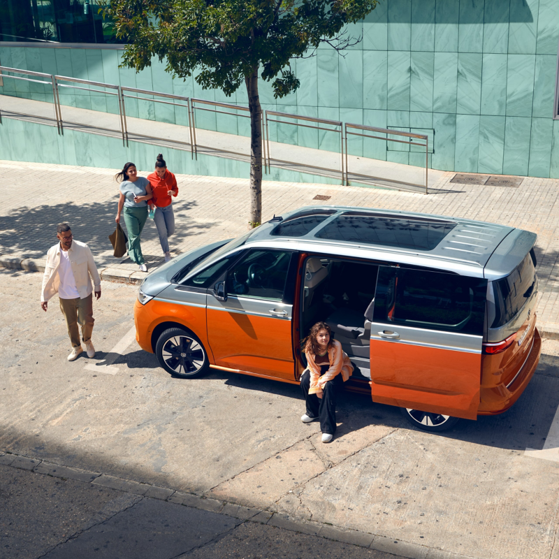 Ein VW Multivan mit offener Schiebetür parkt am Straßenrand und ein menschen laufen darauf zu.