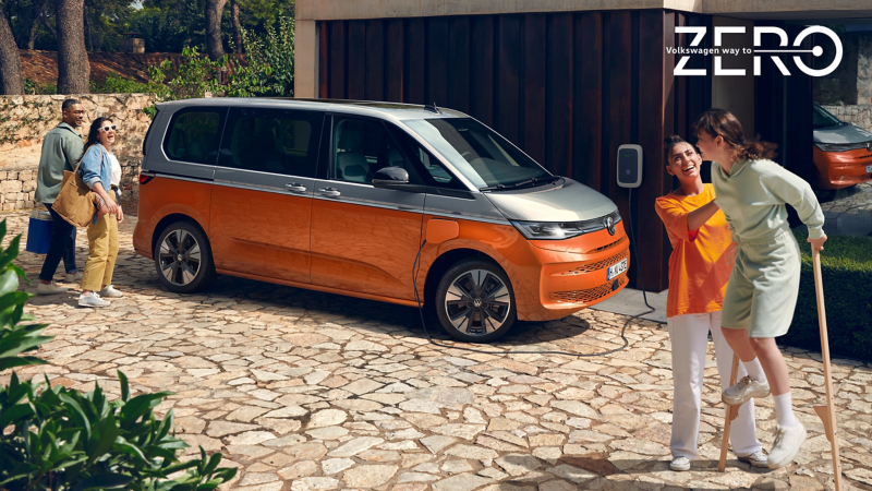Ein VW Multivan lädt an einer Ladestation an einem Haus umgeben von Leuten.