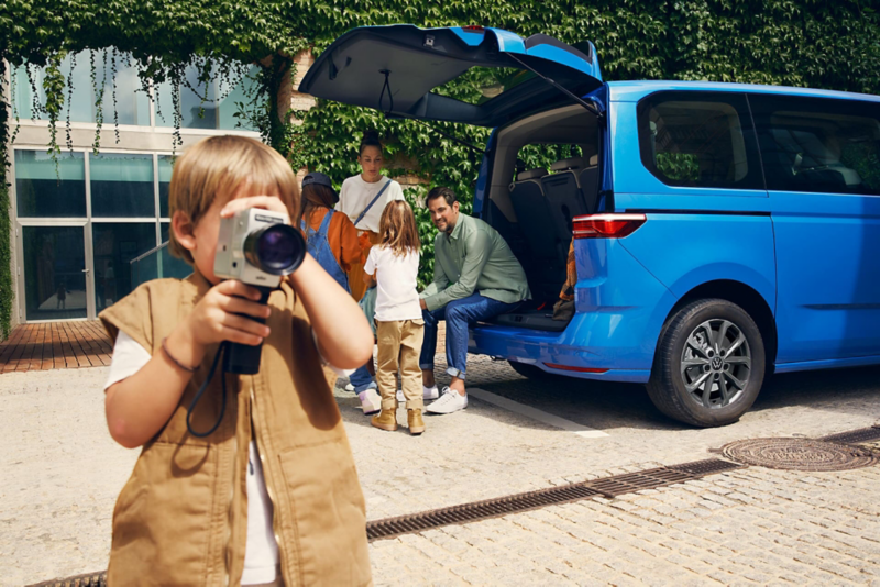 Chłopiec z małą przenośną kamerą na tle rodziny stojącej przy Volkswagenie Multivanie Family z otwartą klapą.