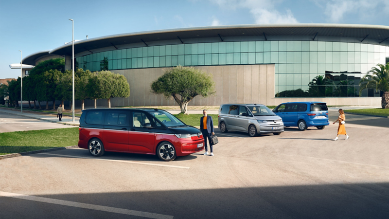  Trois Volkswagen Multivan sont garés sur un parking ensoleillé devant un bâtiment moderne. Un homme en costume sombre se tient près du Multivan rouge et noir au premier plan. Un Multivan gris est garé au milieu, et un Multivan bleu à l'arrière-plan, près d'une femme.