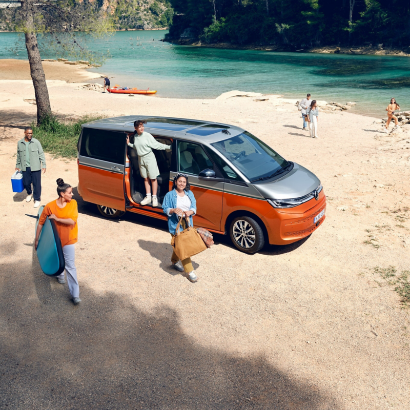 Group of people unloading bags and equipment from a Volkswagen Multivan parked by a beach.