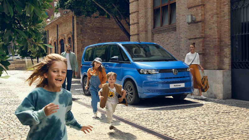 Children running ahead of a Volkswagen Multivan parked on a cobbled street, with adults following behind.