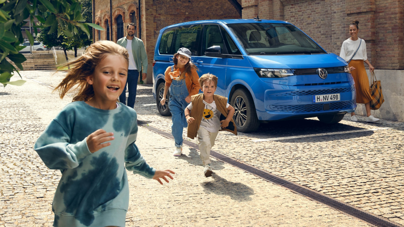 Ein Mann steht neben einem VW Multivan in Medium Blue Metallic und beobachtet Kinder sie Straße hoch laufend.