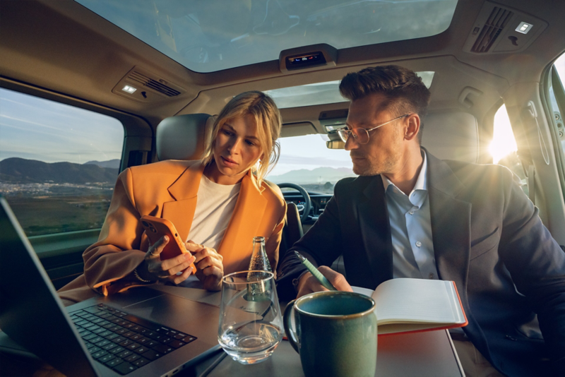 A woman and a man sit on the rear bench seat of a VW and look at a smartphone.