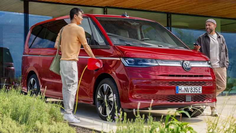 Ein roter VW Multivan eHybrid wird auf einem Hof geladen und zwei Männer stehen daneben