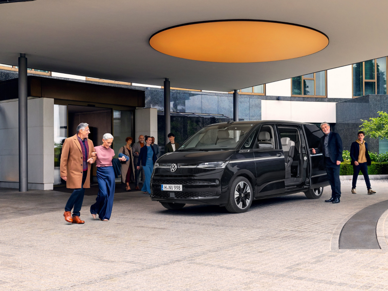 Un Volkswagen Multivan noir est garé sous le porche d'un hôtel moderne, où il sert de navette. Un chauffeur tient la porte coulissante ouverte tandis qu'un groupe de passagers descendus du véhicule se dirige vers l'entrée du bâtiment.