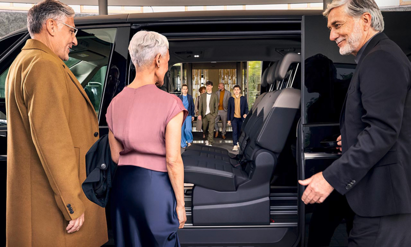 Un chauffeur accueille un couple de passagers à bord d'un Volkswagen Multivan noir. La grande porte coulissante est ouverte, révélant un intérieur spacieux avec des sièges modulables qui facilitent l'accès.