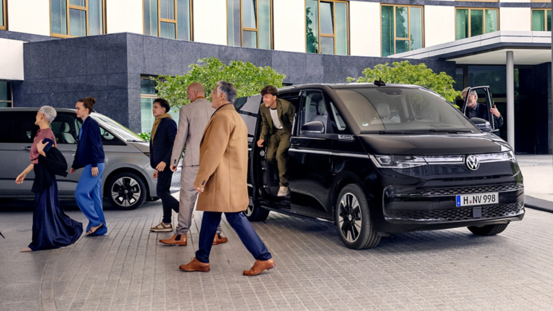 Devant un bâtiment moderne, un Volkswagen Multivan noir sert de navette pour un grand groupe de personnes. Plusieurs passagers se dirigent vers l'entrée tandis qu'un autre sort encore du véhicule, dont le chauffeur tient la porte ouverte.