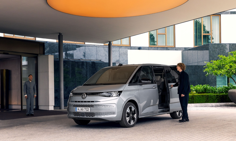 Un Volkswagen Multivan argenté est stationné sous le porche d'un hôtel moderne. Un homme ouvre la porte coulissante du van, tandis qu'un portier se tient à l'entrée du bâtiment.
