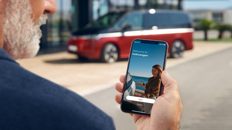 Ein älterer Mann hält ein Smartphone mit der Volkswagen-Anmeldeseite vor einem unscharf im Hintergrund parkenden, rot-schwarzen Van.
