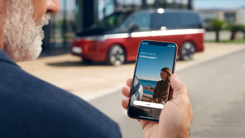 Person holding a smartphone showing the Volkswagen app login screen, with a Volkswagen vehicle parked in the background.
