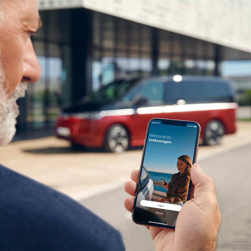 Ein Mann hat die Volkswagen App auf seinem Smartphone geöffnet, im Hintergrund steht ein Multivan.