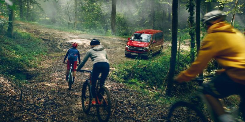 Trzy osoby jadą na rowerach w lesie. W tle czerwony Volkswagen Multivan 6.1.
