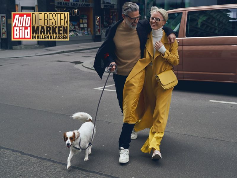 Ein Mann und eine Frau mit einem Hund machen einen Spaziergang. Im Hintergrund steht ein Multivan. In der oberen linken Ecke findet sich das Logo des Auto Bild Awards zur Marke des Jahres.