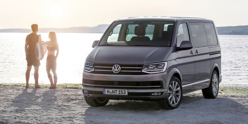 Et par surfere står på stranden ved siden af Multivan fra Volkswagen Erhvervsbiler.