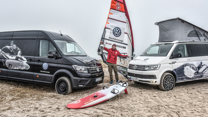 Vincent Langer står med sit surfbræt mellem to Multivan-biler på stranden.