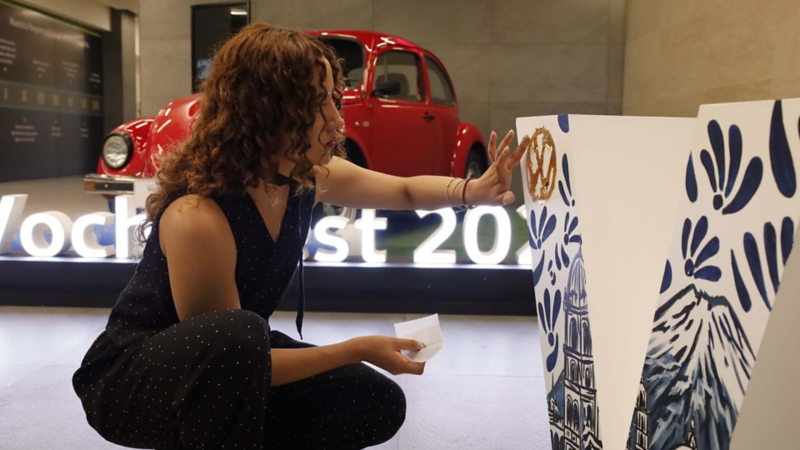 Mujer decora letra gigante con patrones azules junto a Volkswagen Sedan de color rojo.