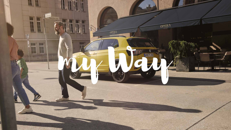 Un Volkswagen de color amarillo parado frente a una cafetería con una familia pasando delante con el logo de My Way
