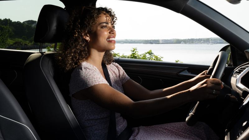 A smiling woman with curly hair drives a Volkswagen, with a view of a harbor in the background.