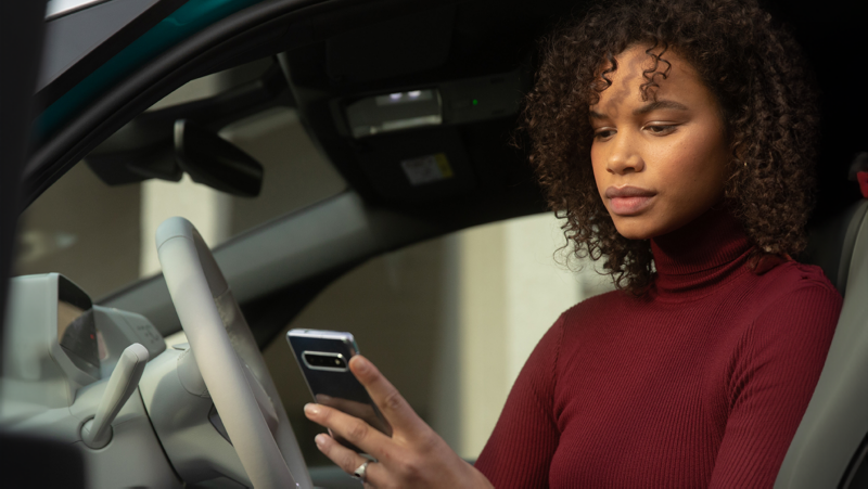 Une femme aux cheveux bouclés regarde son téléphone tout en étant assise sur le siège conducteur d'une voiture.