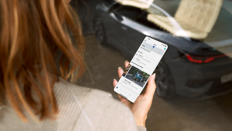 A person holds a smartphone displaying a myVW app with options for viewing driving data and the owner's manual.