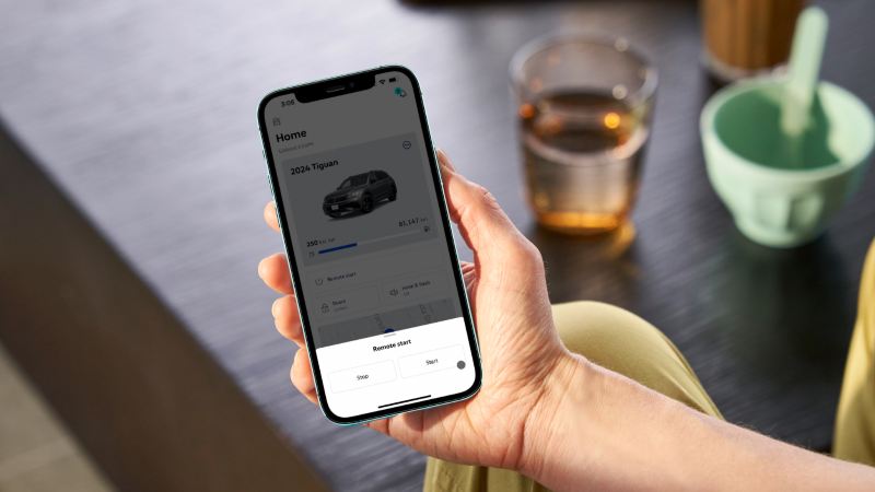 Une personne tient un téléphone affichant une interface myVW pour une voiture, avec une fenêtre contextuelle "Démarrage à distance" ouverte.