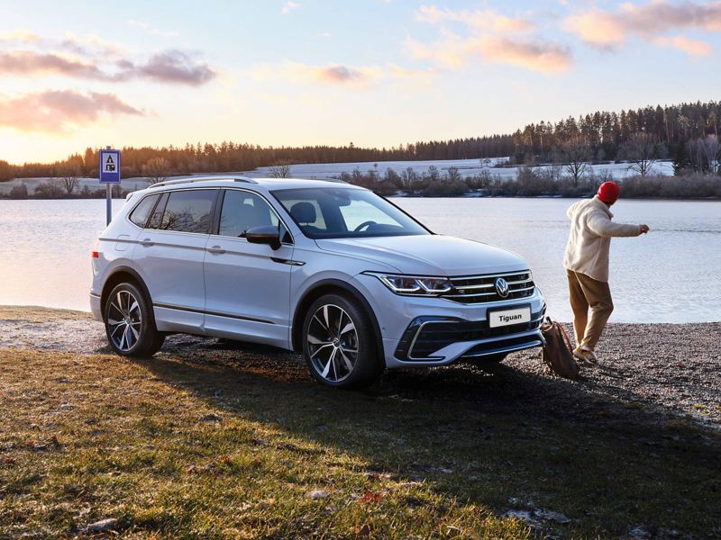A white Tiguan Allspace R-Line with tinted rear windows and switched-on light signature stands on the lakeshore.