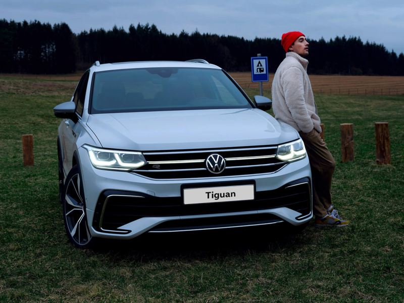 A man leans against the hood of a white Tiguan Allspace R-Line, the LED matrix headlights and light bar on. 