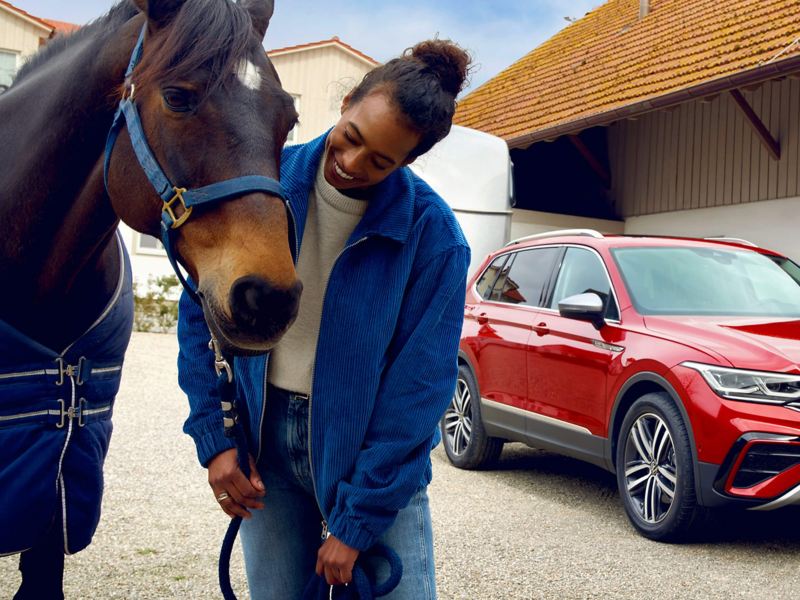 A red Tiguan Allspace Elegance with a horse trailer is parked at a horse farm. A woman with a brown horse is standing in front of it. 
