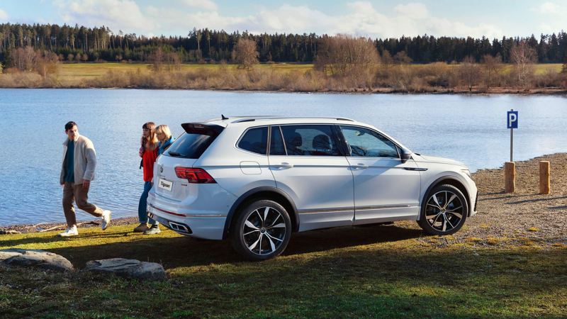 A group of young people walk past a white Tiguan Allspace R-Line parked by a lake.